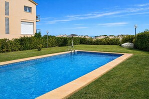 Una piscina rectangular con borde de azulejos, rodeada por un césped cuidado y un edificio de fachada color crema.