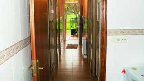 Un pasillo con paredes de baldosas, una puerta de madera y vista a un jardín por una puerta de vidrio.