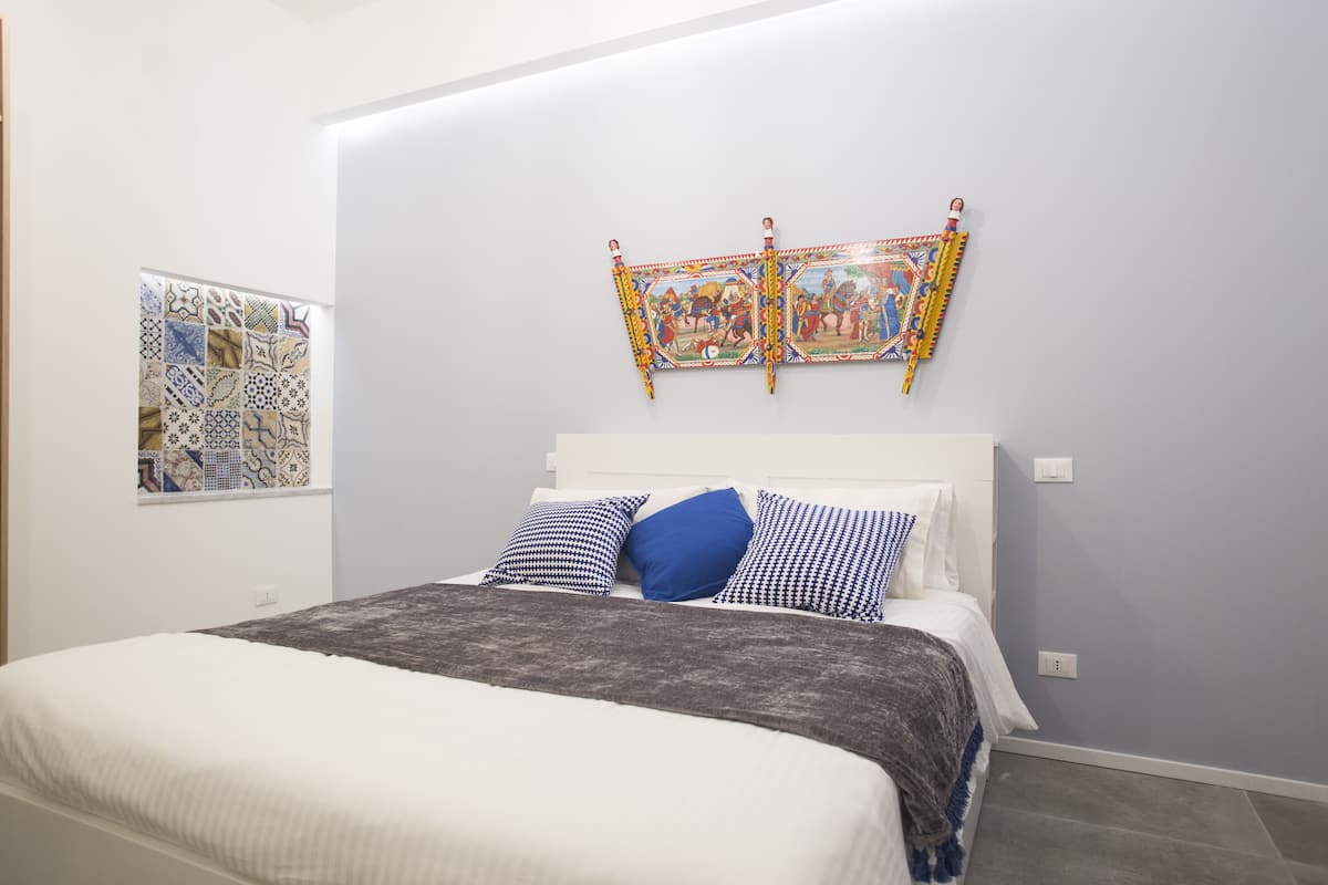 A bedroom with a bed, pillows, a blanket, a wall-mounted artwork, and a decorative tile wall.