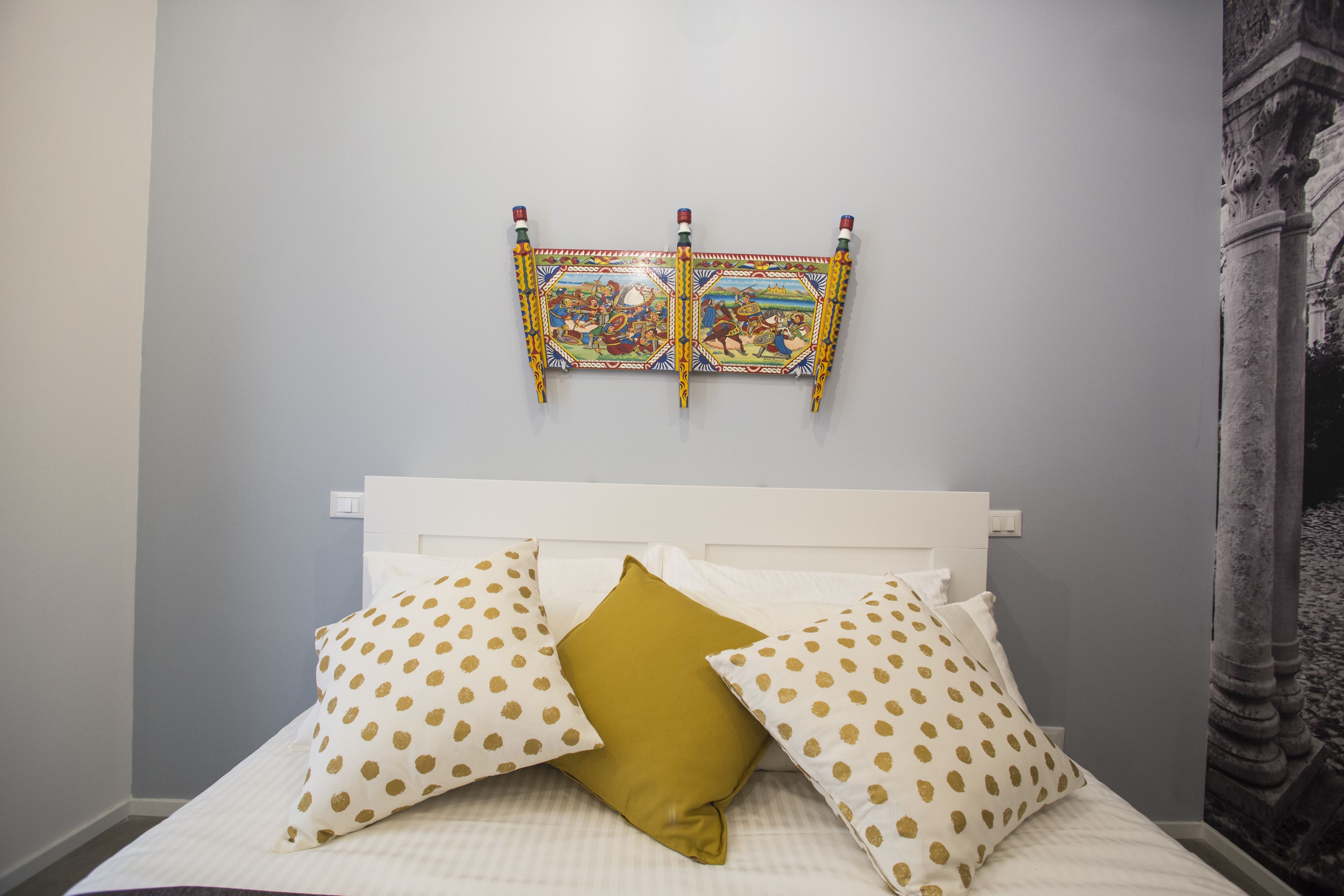 A bed with white bedding and gold polka dot pillows, a white headboard, and a colorful tapestry on the wall.