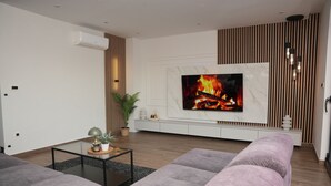 Uma sala de estar moderna com televisão de tela plana presa a uma parede branca, sofá cinza, mesa de centro de vidro e piso de madeira.