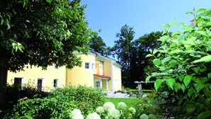 A yellow building with a red roof, surrounded by lush greenery and white flowers.