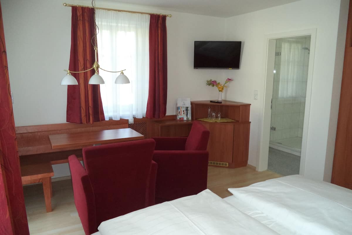 Quarto de hotel com mesa de jantar de madeira, cadeiras vermelhas, cama com roupa de cama branca, televisão fixada na parede e área de chuveiro.