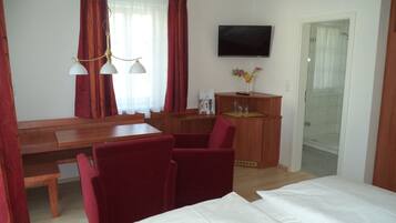 Ein Hotelzimmer mit Holztisch, roten Stühlen, einem Bett mit weißer Bettwäsche, einem an der Wand montierten Fernseher und einer Duschkabine.