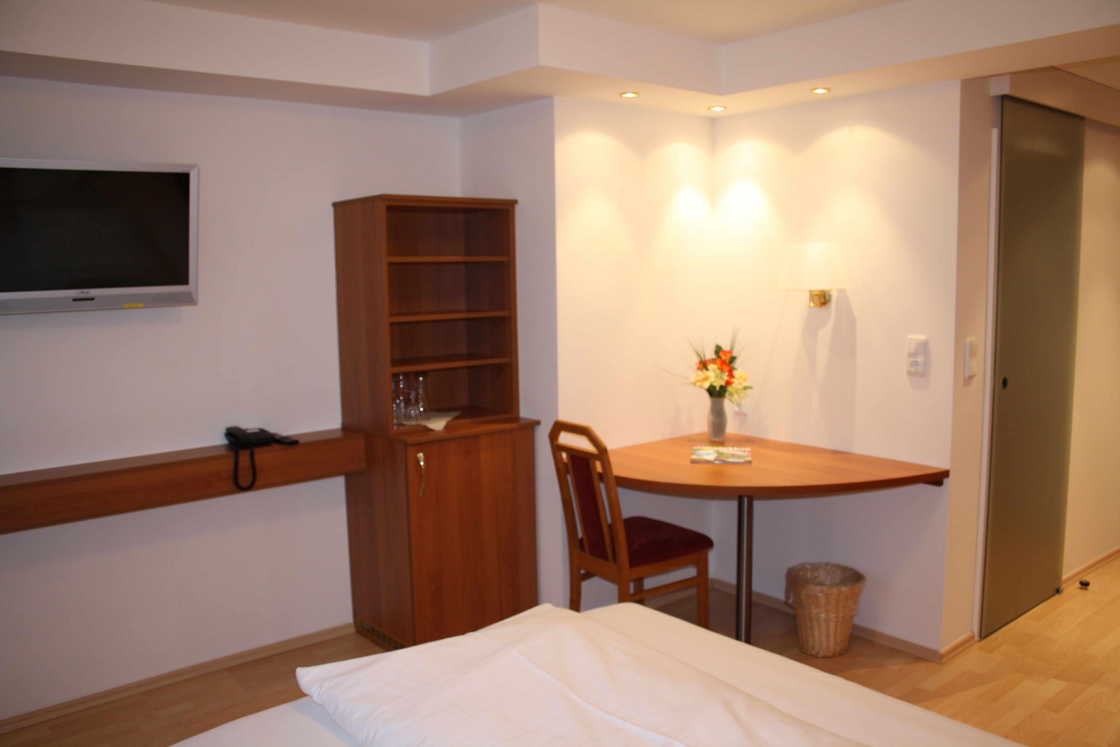 A hotel room with a bed, a wooden cabinet, a television mounted on the wall, a small table with a chair, and a vase with flowers.