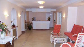 A hotel lobby with a reception desk, a dining area with wicker chairs and a glass table, and a decorative vase with flowers.