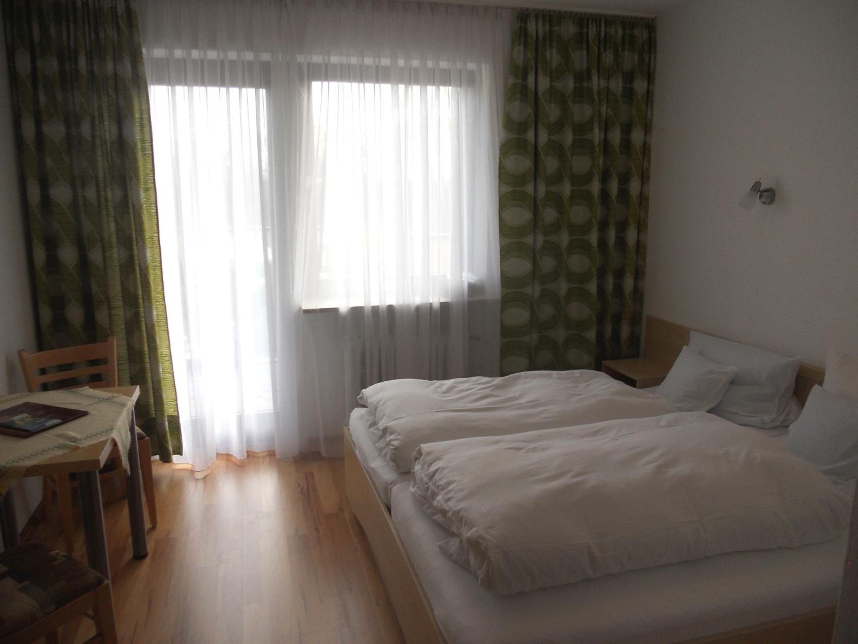 A hotel room with two beds, a wooden table with a chair, and sheer curtains.