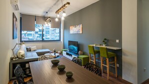 A modern living room with a sofa, dining table, and chairs, a TV, and a large window with a city view.