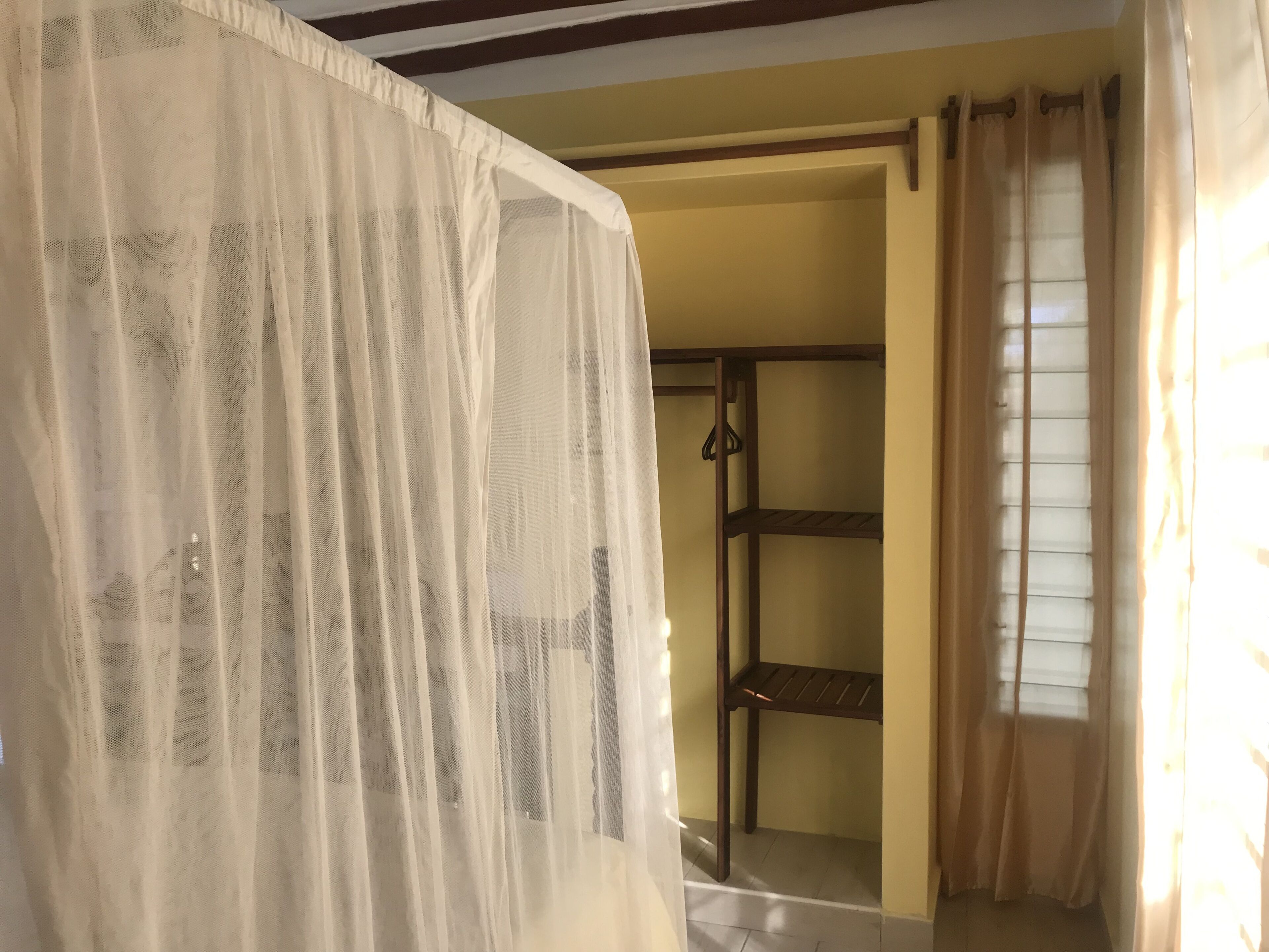 A room with a bed covered by a sheer white curtain, a wooden shelf unit, and a window with curtains.