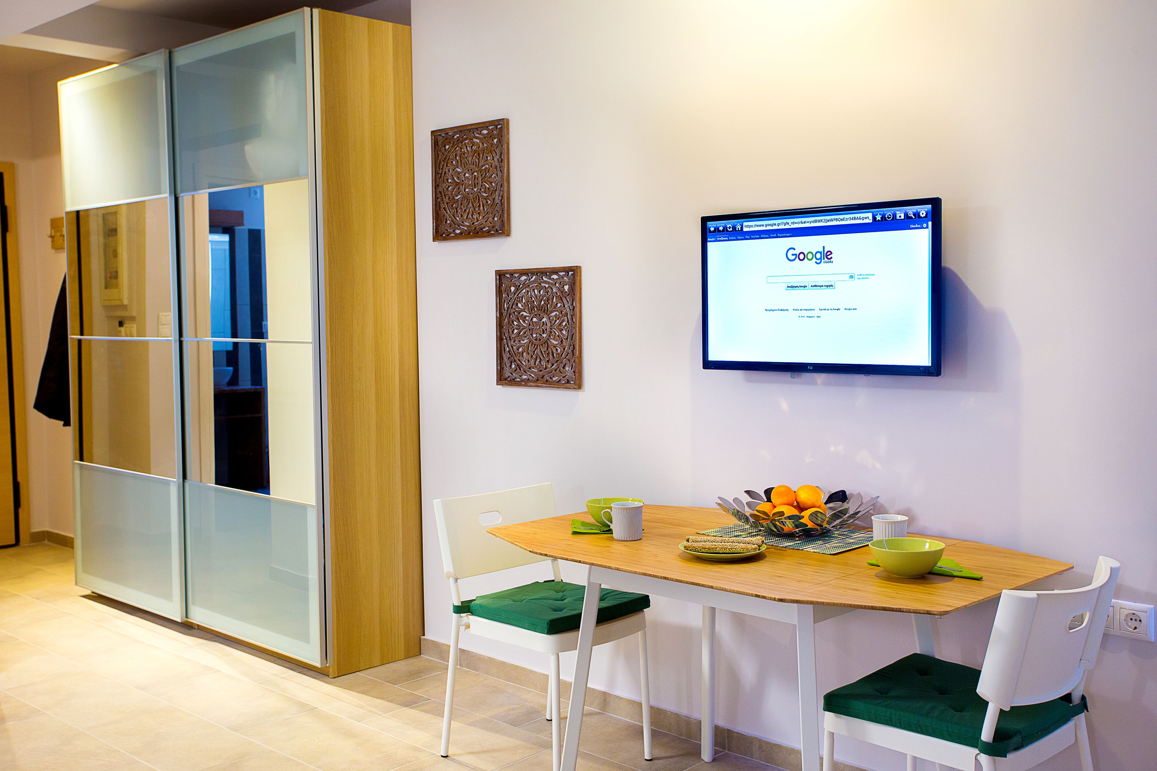 A modern dining area with a wooden table, white chairs, and a green cushion. A television mounted on the wall displays the Google homepage.
