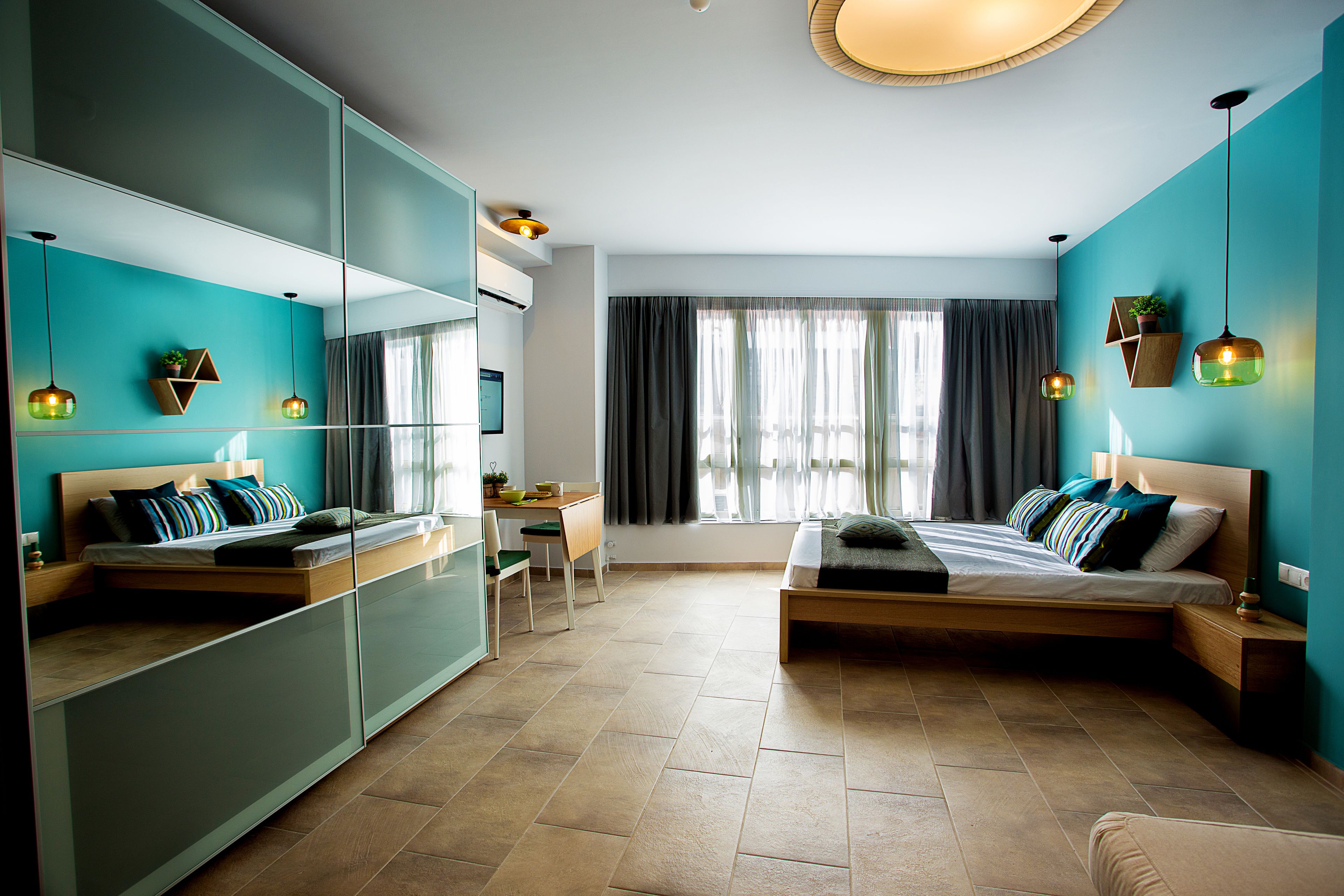 A modern bedroom with a bed, a desk, and a chair. There is a large mirror and a window with curtains.