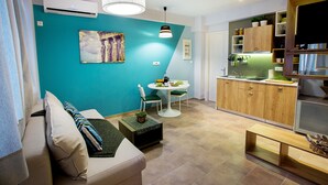 A compact living space with a teal accent wall, a sofa with striped cushions, a small dining table, and a kitchenette area. - Emerald Suite (Athens)