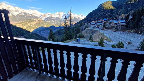 A balcony with a view of a mountain range, a parking area, and buildings with red roofs.