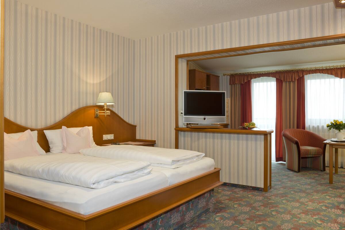 A hotel room with a large bed, a television on a stand, a desk with a chair, and a view through an open door.