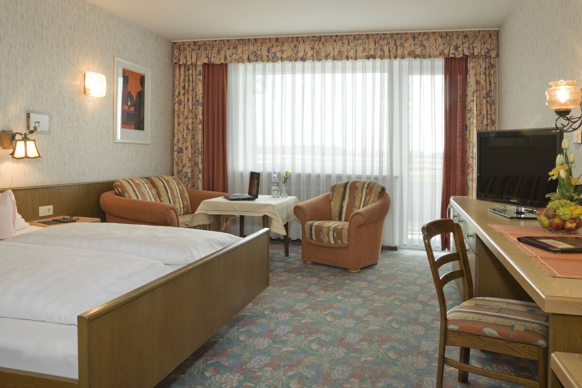A hotel room with a bed, a desk with a television, a chair, a small table, and a window with curtains.