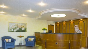 Un área de recepción con un mostrador de madera, dos sillones azules, un cuadro de flores y el símbolo de accesibilidad para sillas de ruedas en el suelo.