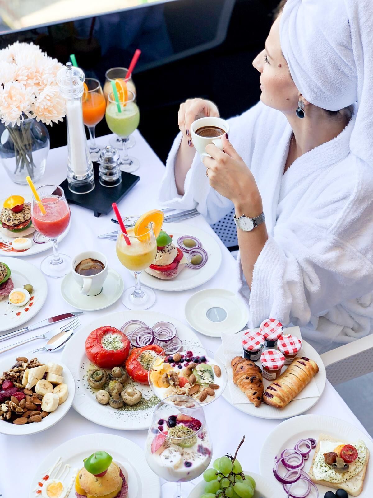 A person in a white robe at a table with various breakfast items, including pastries, fruits, and drinks.