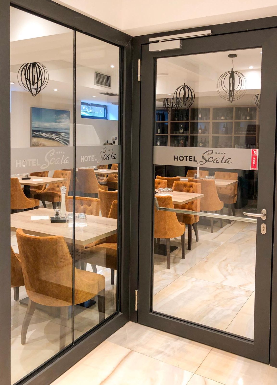 Un interior de hotel moderno con puertas de vidrio, una zona de comedor con mesas y sillas, y un bar visible con estantes llenos de botellas.