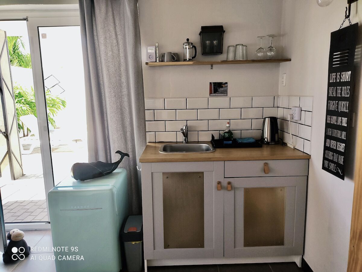 Une cuisine compacte équipée d’un évier, d’une machine à café et d’une bouilloire sur le plan de travail. On y trouve également un petit réfrigérateur et un placard doté d’une porte en treillis métallique. Un poster affichant une citation motivante est visible sur le mur.