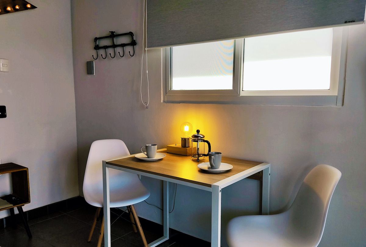 A small dining area with a table, two chairs, a coffee maker, and a window with blinds.