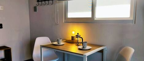 A small dining area with a table, two chairs, a coffee maker, and a window with blinds.