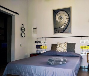 Une chambre à coucher avec un lit, des oreillers, un luminaire fixé au mur et un tableau représentant un escalier en colimaçon.