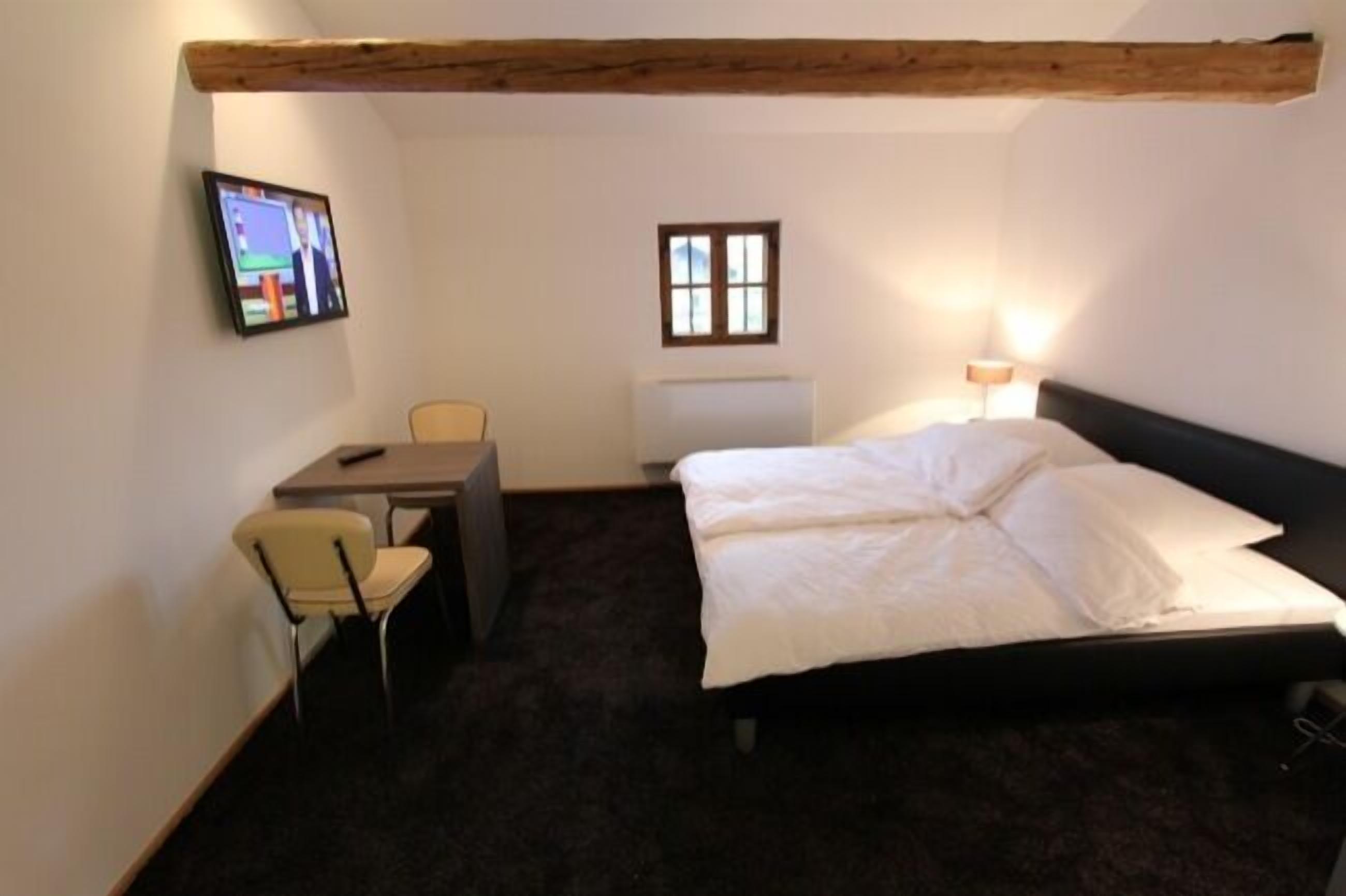 A small, cozy room with a bed, a desk, a chair, a television, and a window.