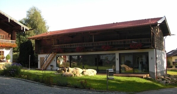 A two-story wooden house with a balcony, large windows, and a stone pathway leading to the entrance.