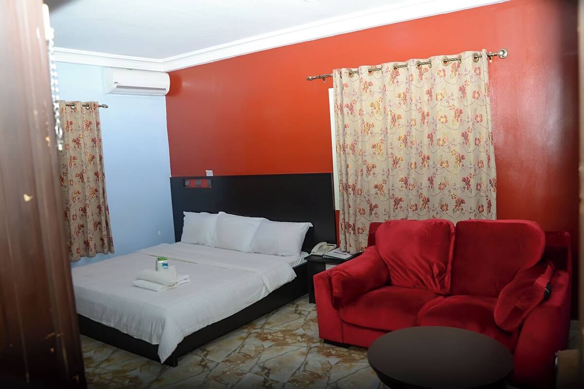 A hotel room with a bed, a red armchair, a small round table, and a nightstand with a phone and a lamp.