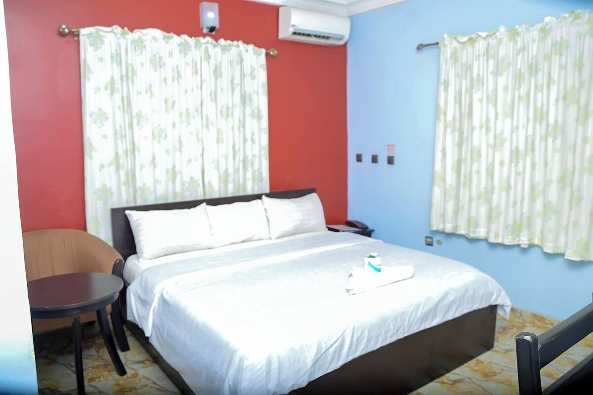 A hotel room with a bed, a chair, a bedside table, and an air conditioning unit.