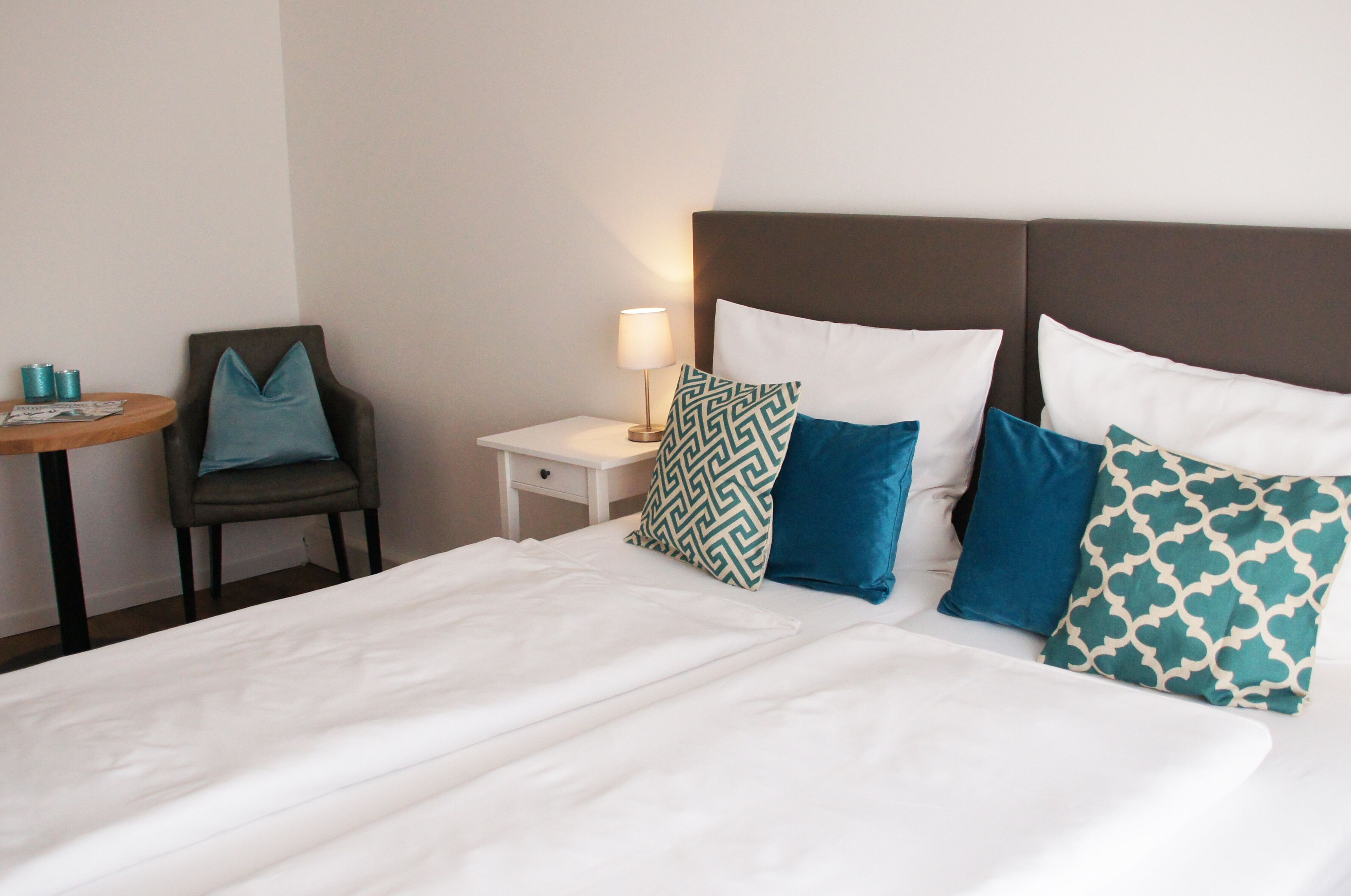A hotel room with a bed, pillows, a nightstand, a lamp, a chair, and a small table.