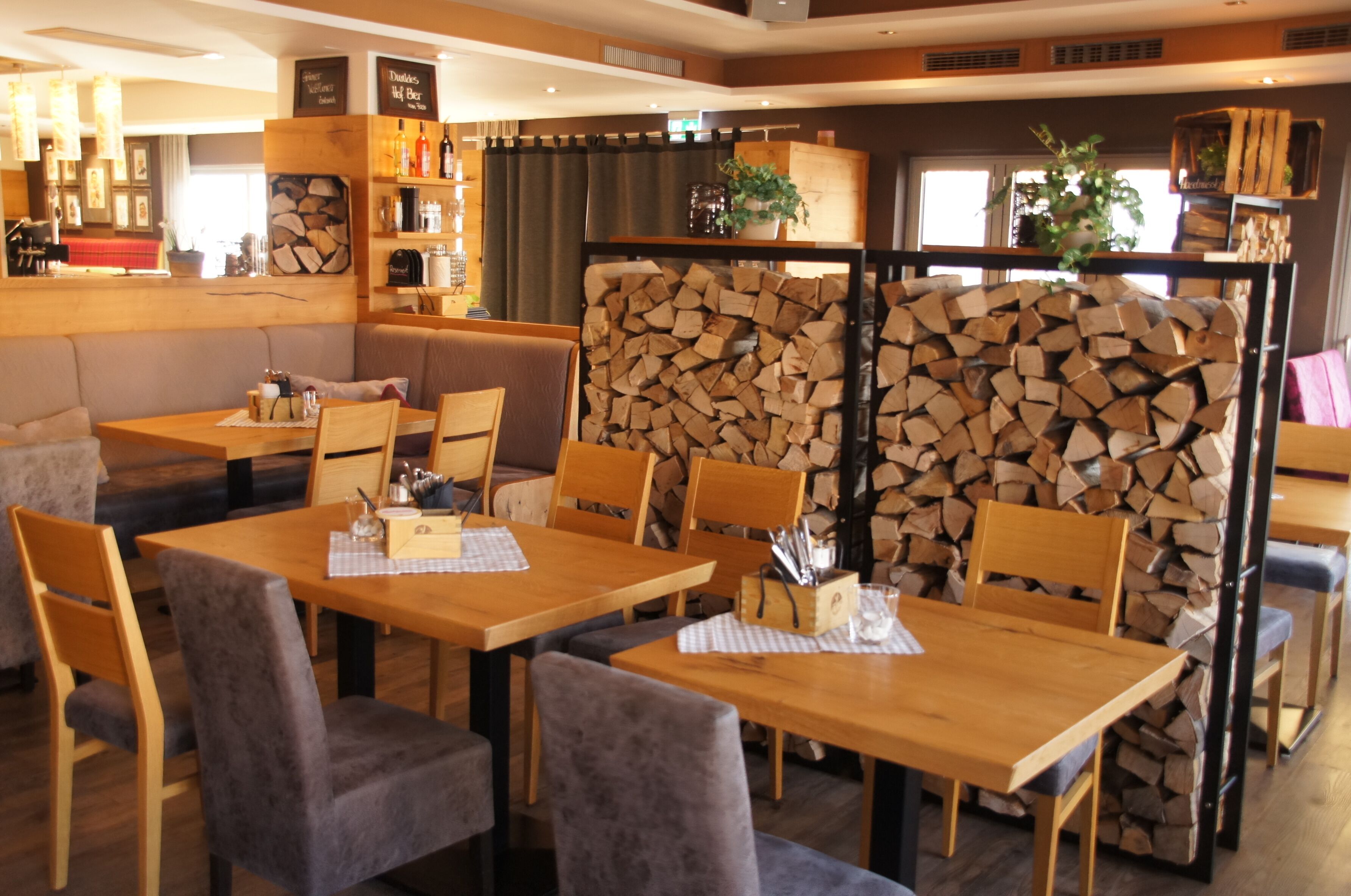 A cozy restaurant with wooden tables and chairs, a stack of firewood in the background, and various kitchen items on shelves.