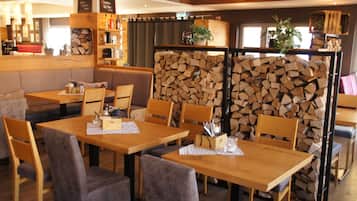 Un restaurante acogedor con mesas y sillas de madera, leña apilada en el fondo y varios utensilios de cocina en estantes.