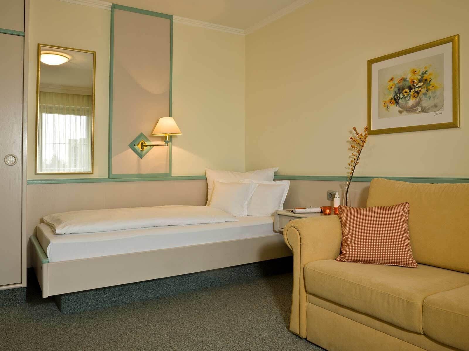 A hotel room with a bed, a bedside table, a lamp, a couch, and a framed picture on the wall.