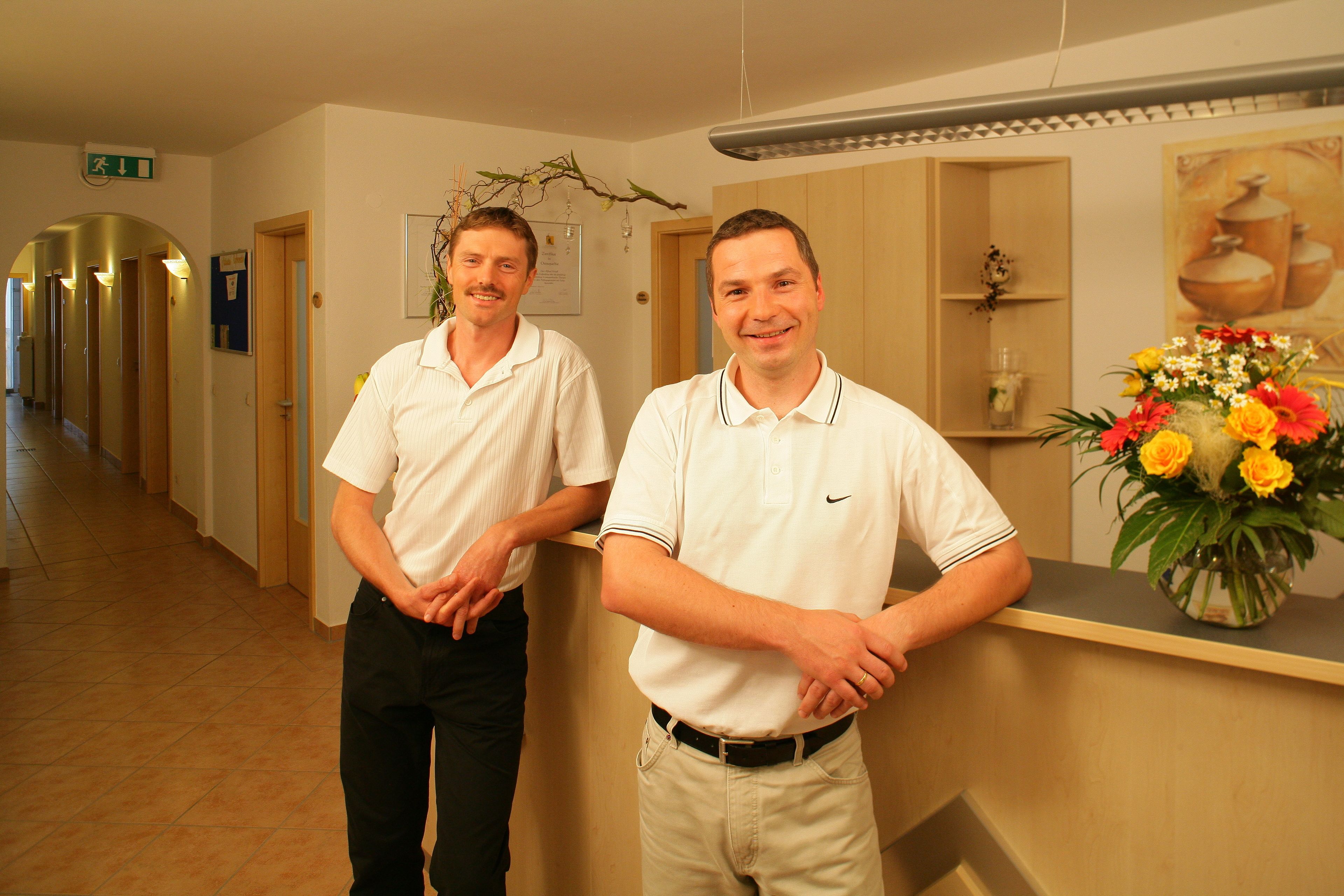 Two men standing in a hallway with a reception desk, a plant, and a door with a sign.