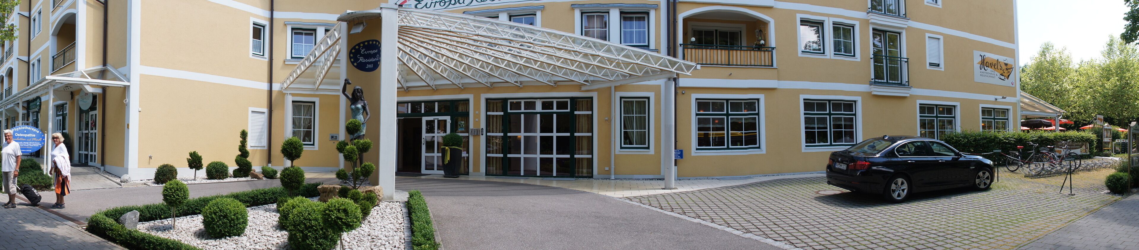 L’entrée d’un hôtel, avec une allée couverte, une voiture noire garée devant et un jardin bien entretenu.