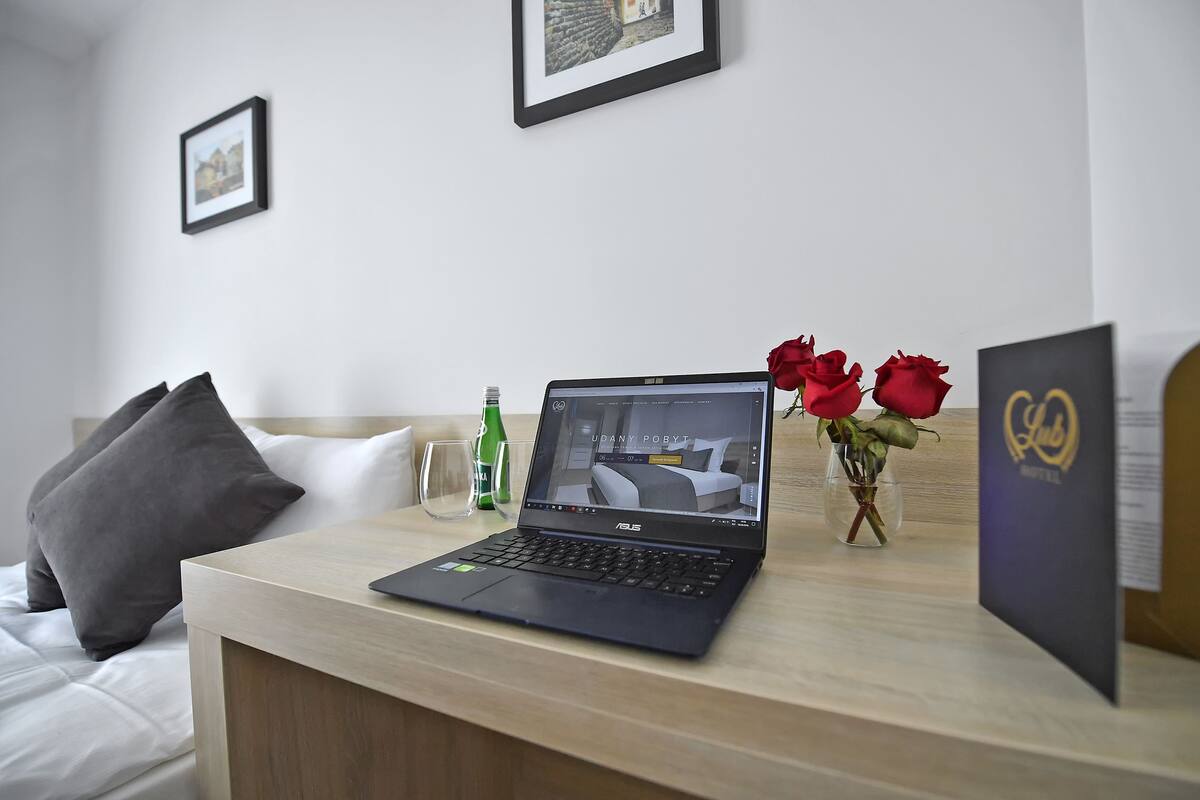 Un ordenador portátil sobre un escritorio de madera en una habitación de hotel con cama, almohadas, un jarrón con rosas rojas y un cuadro en la pared.
