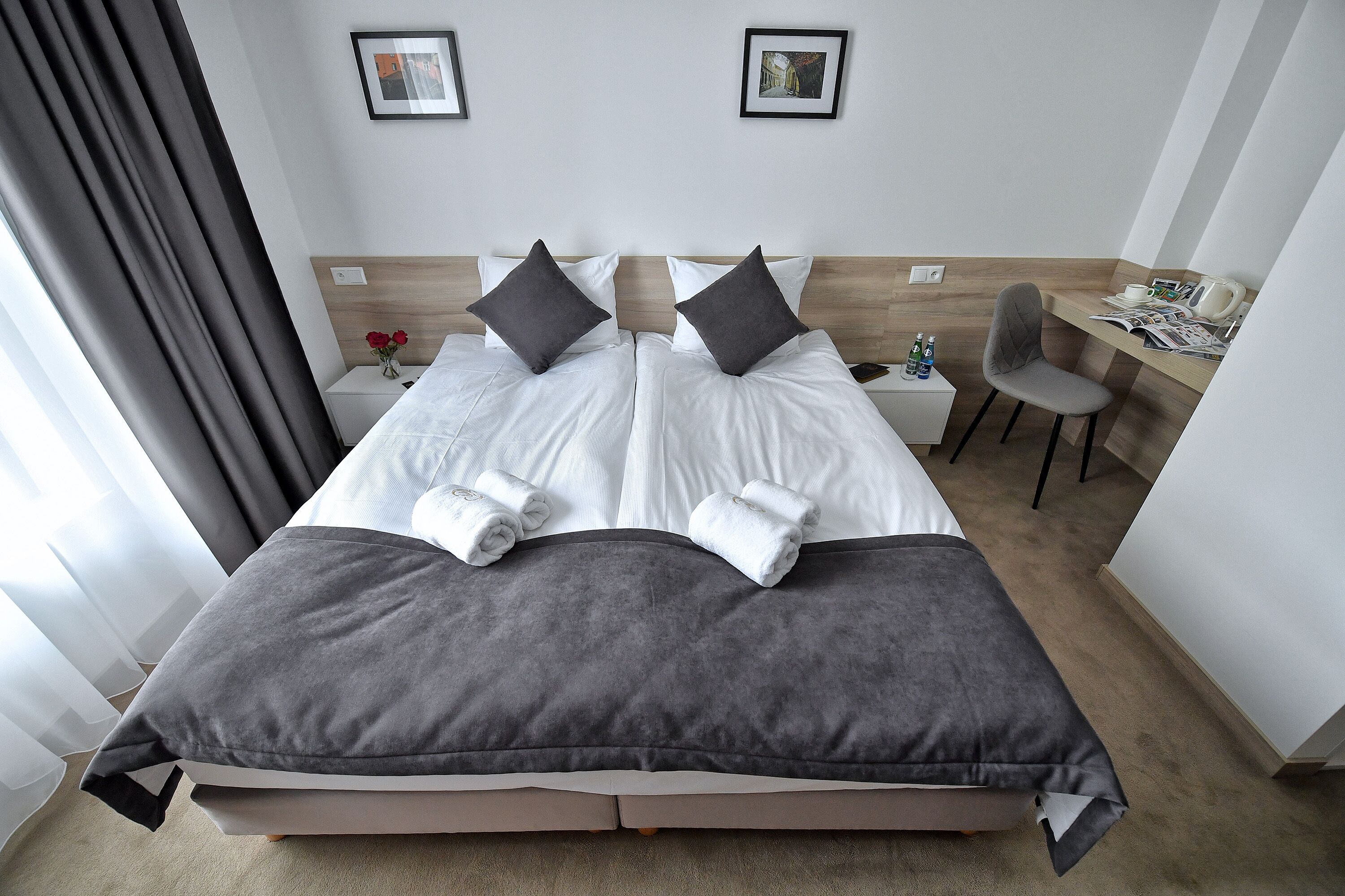 A hotel room with a double bed, two pillows, a desk with a chair, and a small table with a vase of flowers.