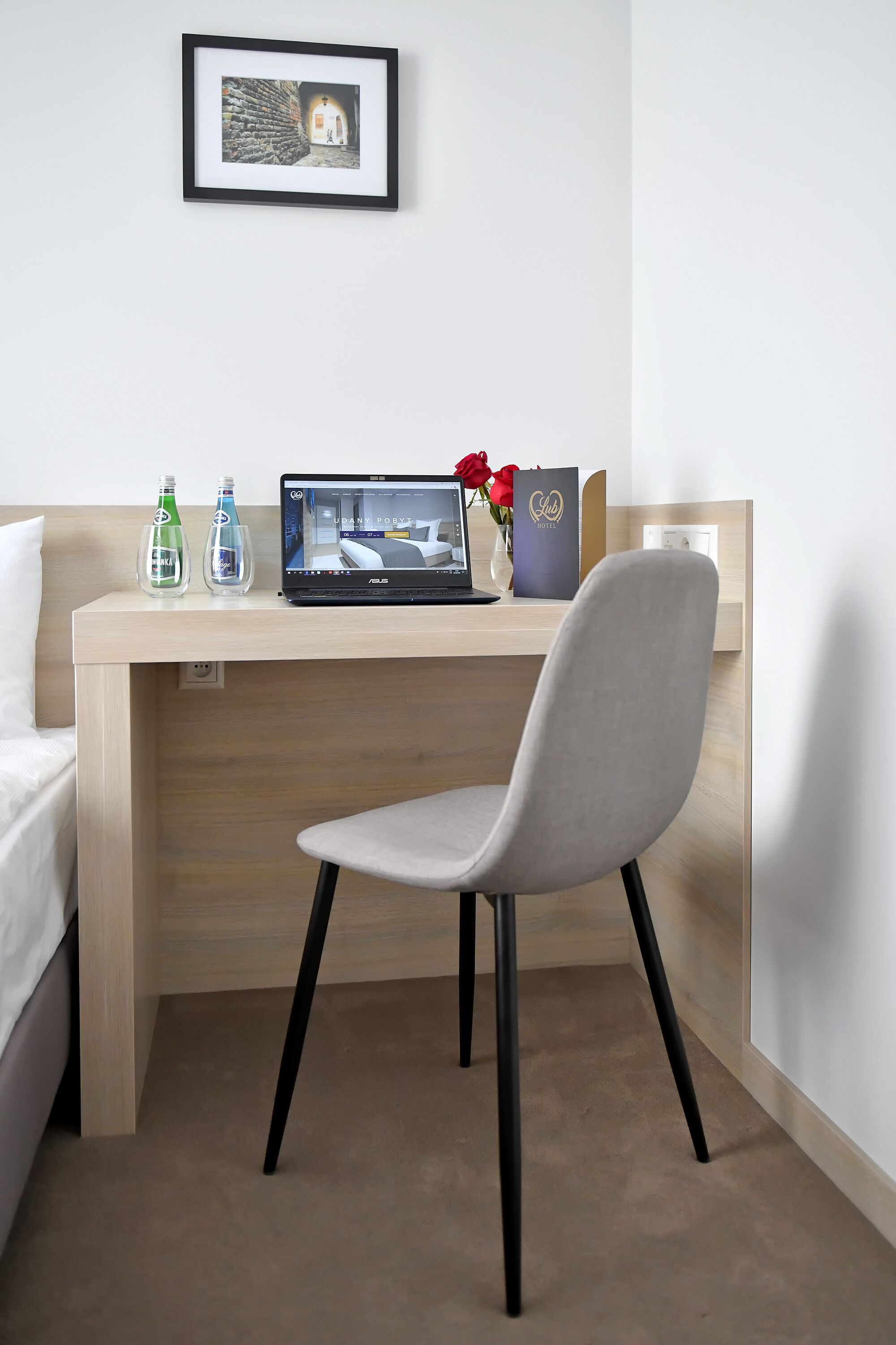 A hotel room with a bed, a desk with a laptop, a chair, and a framed picture on the wall.