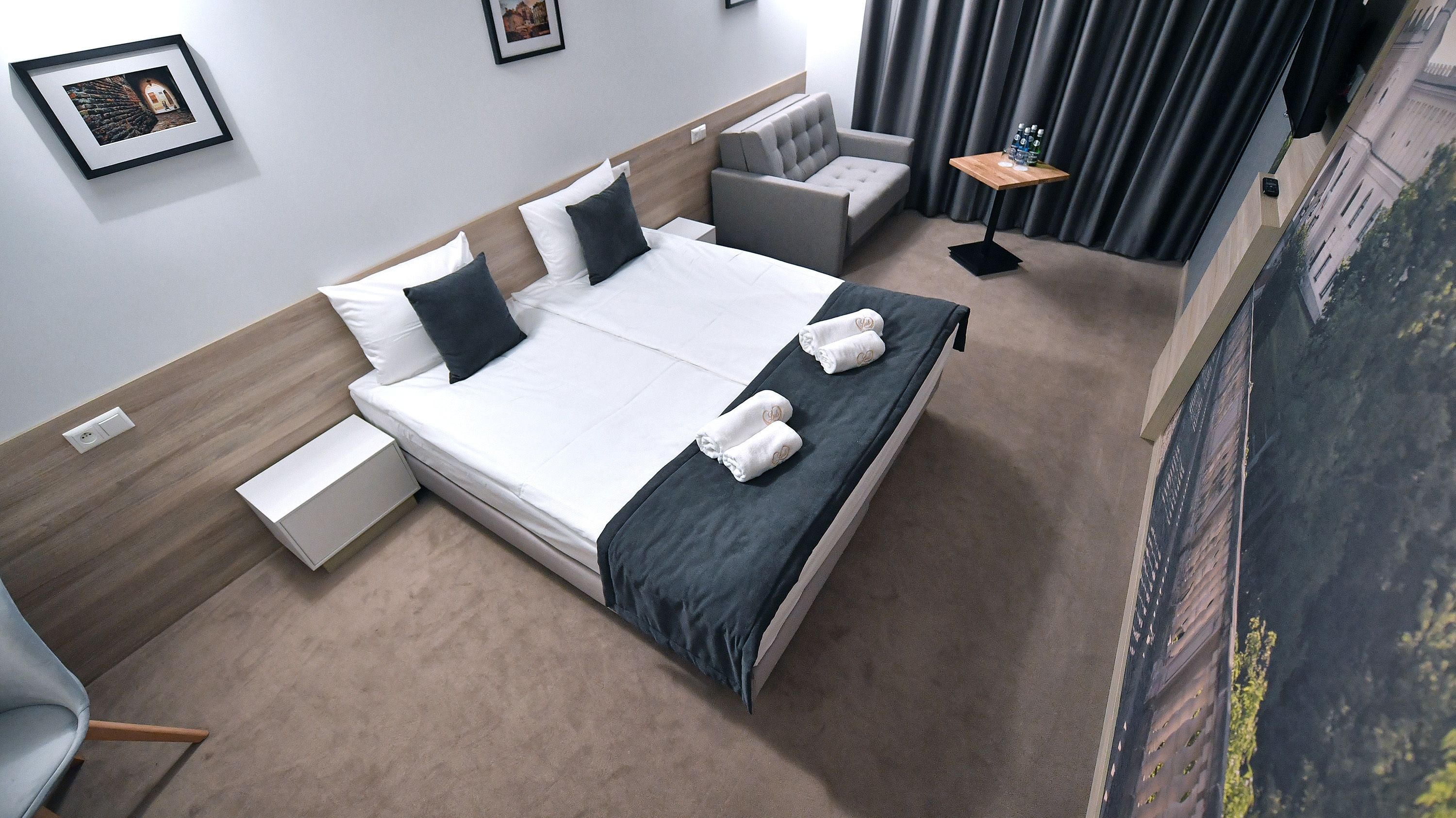 A modern hotel room with a large bed, a grey sofa, a small table with bottles, and a painting on the wall.