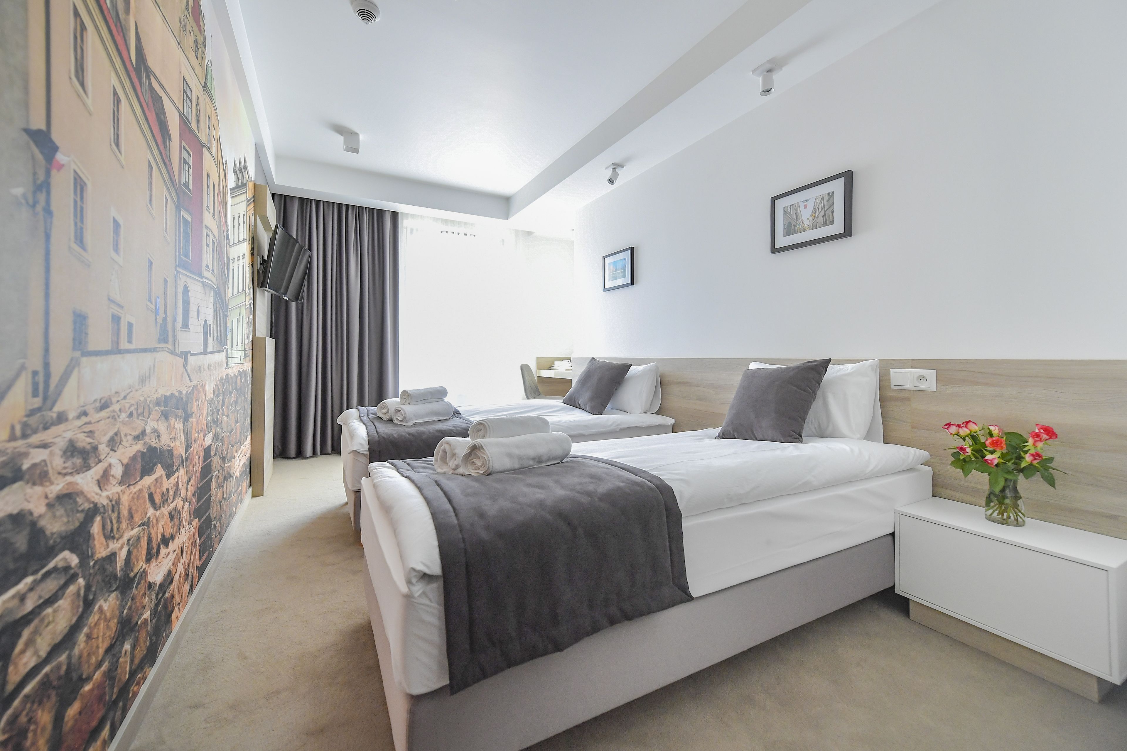 A hotel room with two beds, a large window, a painting on the wall, and a vase with flowers on the nightstand.