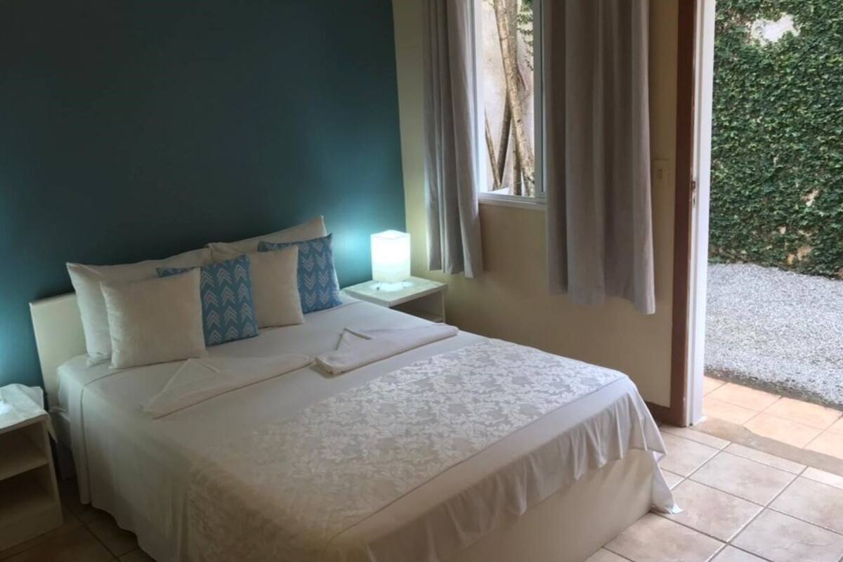 Un lit bien fait, avec du linge de lit blanc et des coussins à motifs bleus, une table de chevet avec une lampe, et une vue sur un jardin à travers une fenêtre ornée de rideaux.