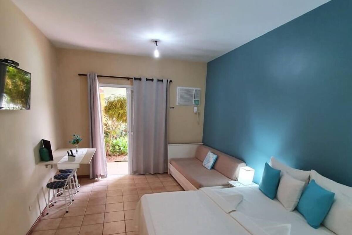 Une chambre d’hôtel avec un lit, un bureau et une chaise. Il y a une fenêtre avec des rideaux, un luminaire au plafond et un climatiseur fixé au mur.