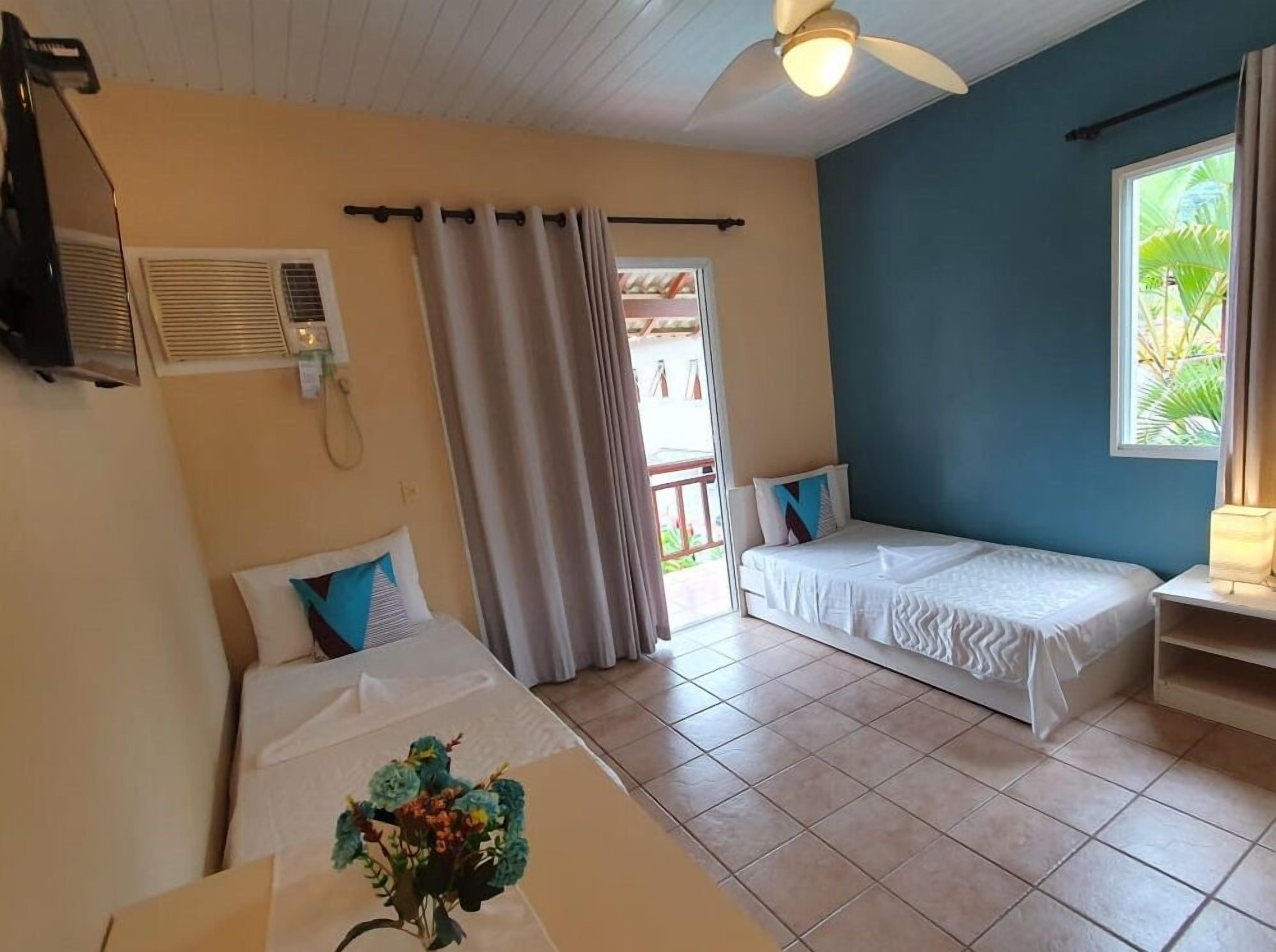 Quarto com duas camas, um ventilador de teto, janela com cortinas e uma mesinha de centro com um vaso.