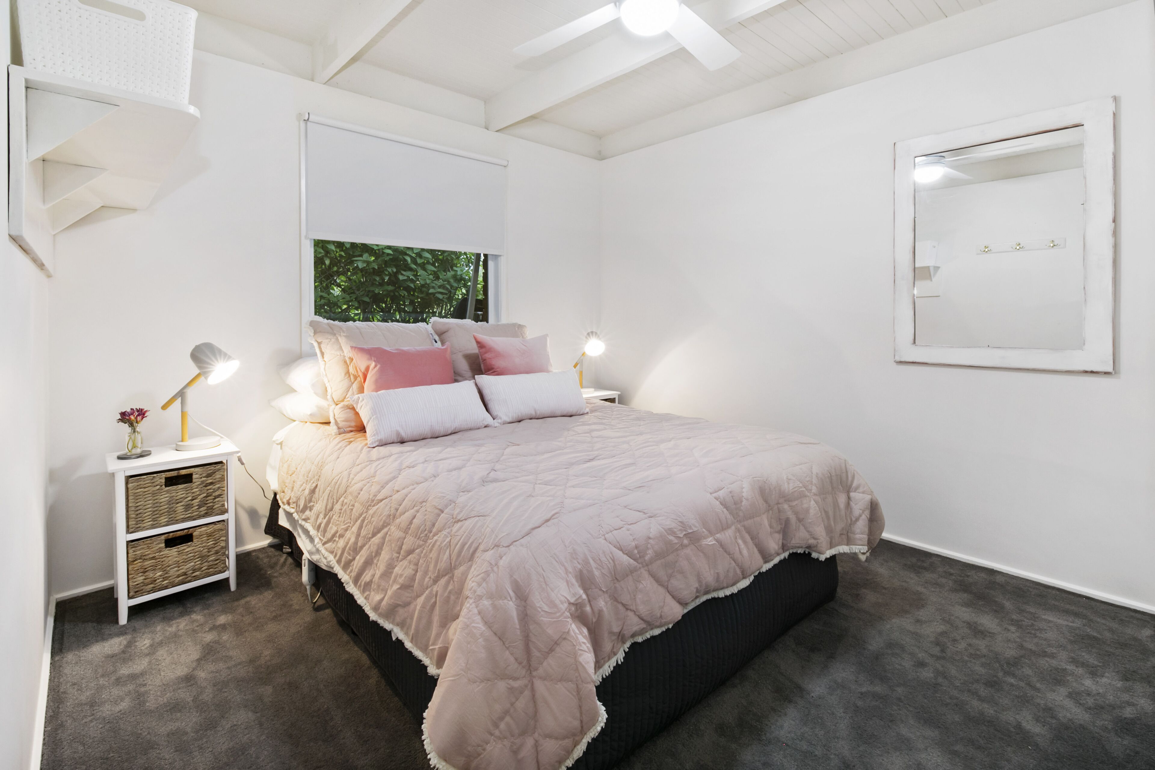 Une chambre à coucher comprenant un lit, des tables de chevet, un miroir fixé au mur et une fenêtre avec des stores.