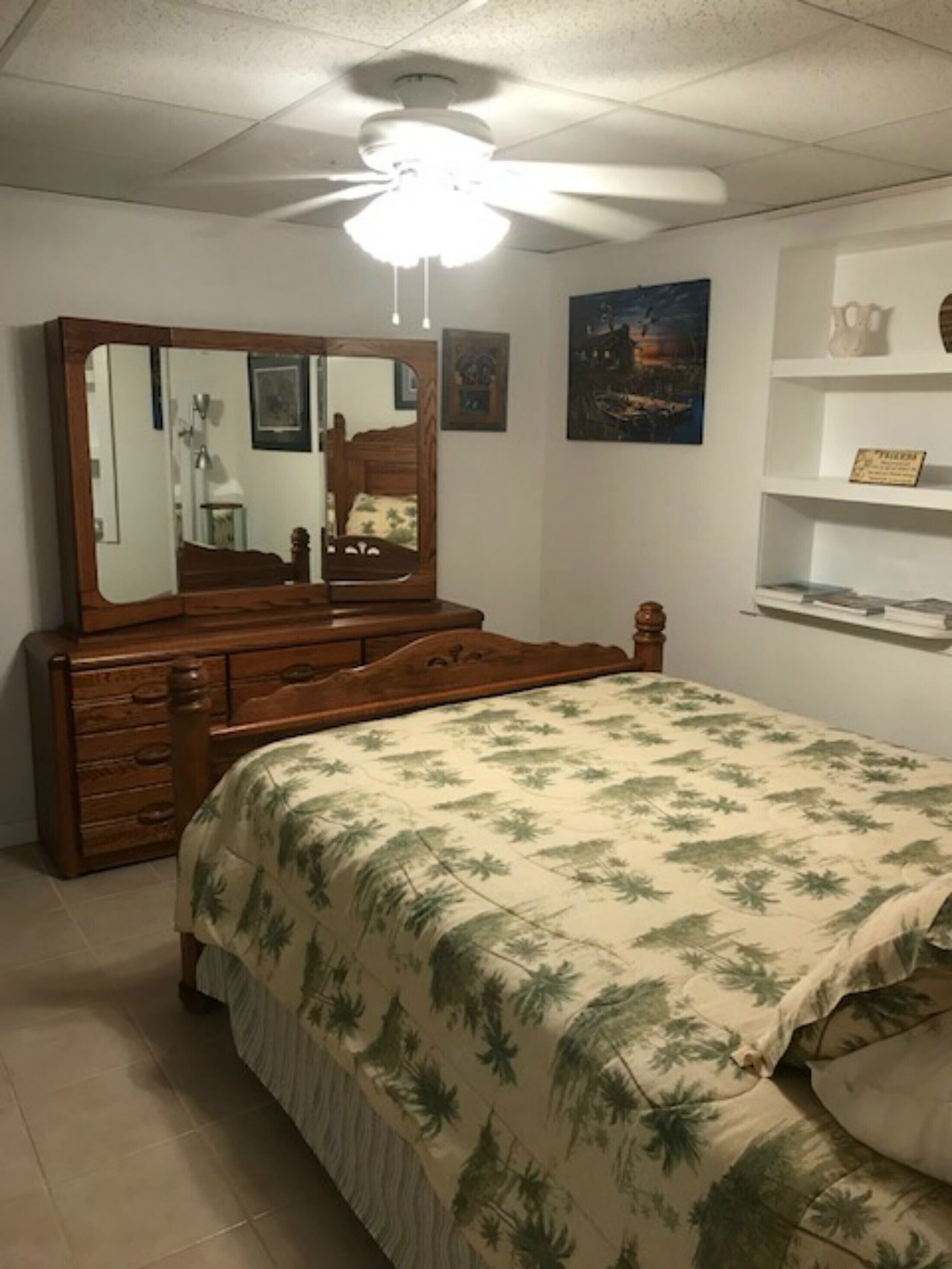 Une chambre à coucher comprenant un lit, une commode, un ventilateur de plafond et une étagère fixée au mur.