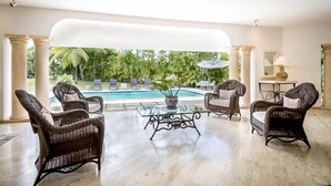 A spacious indoor area with wicker furniture, a glass coffee table, and a view of a swimming pool and greenery outside. - Casa de Campo, Beautiful Villa with Pool Next to Golf Course and Beach/Sleeps 8 (La Romana)