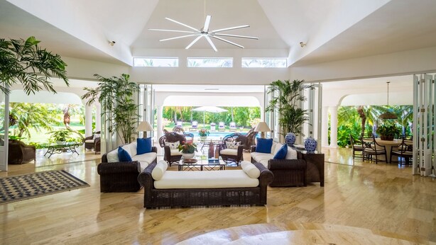 A spacious indoor area with a central seating area featuring wicker sofas and a coffee table, surrounded by large windows offering views of a pool and lush greenery. - Casa de Campo, Beautiful Villa with Pool Next to Golf Course and Beach/Sleeps 8 (La Romana)