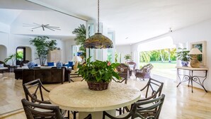 A spacious living area with a round table surrounded by wicker chairs, a hanging light fixture, and a view of a lush garden through large windows. - Casa de Campo, Beautiful Villa with Pool Next to Golf Course and Beach/Sleeps 8 (La Romana)