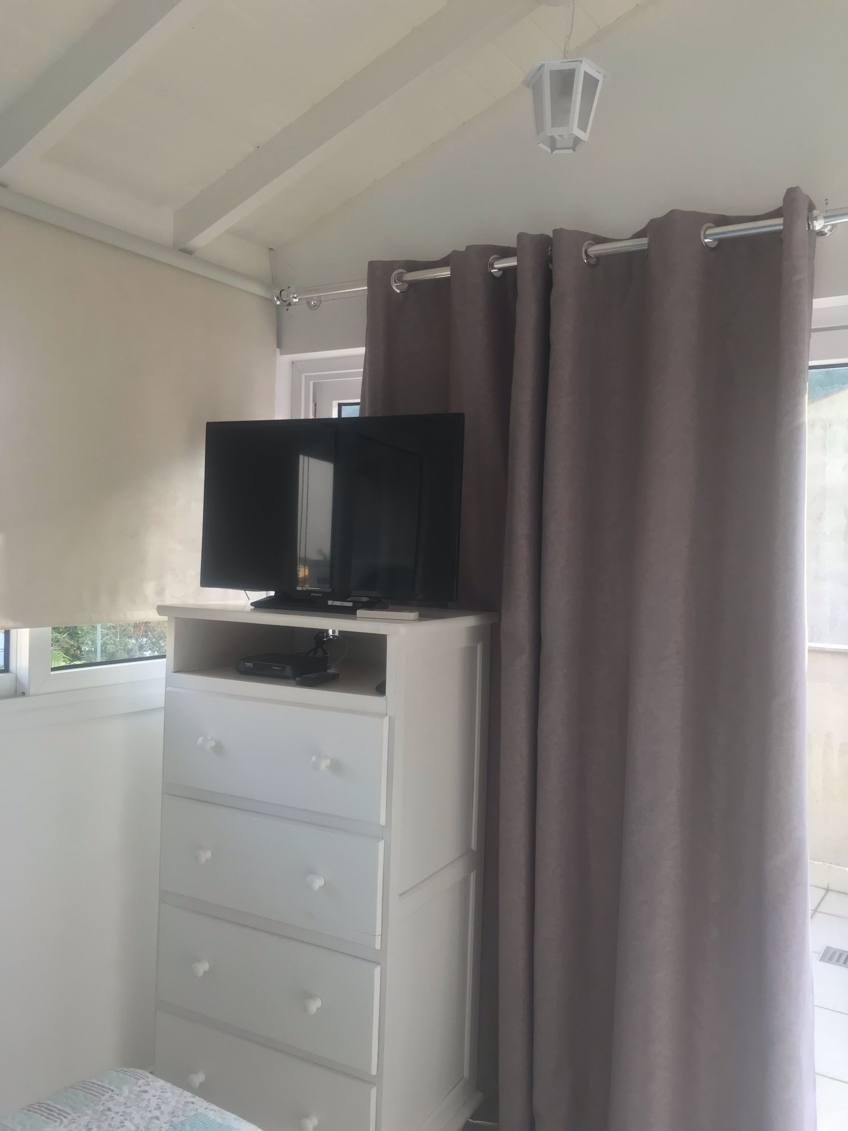 Een slaapkamer met een televisie op een dressoir, een raam met gordijnen en een plafondlamp.